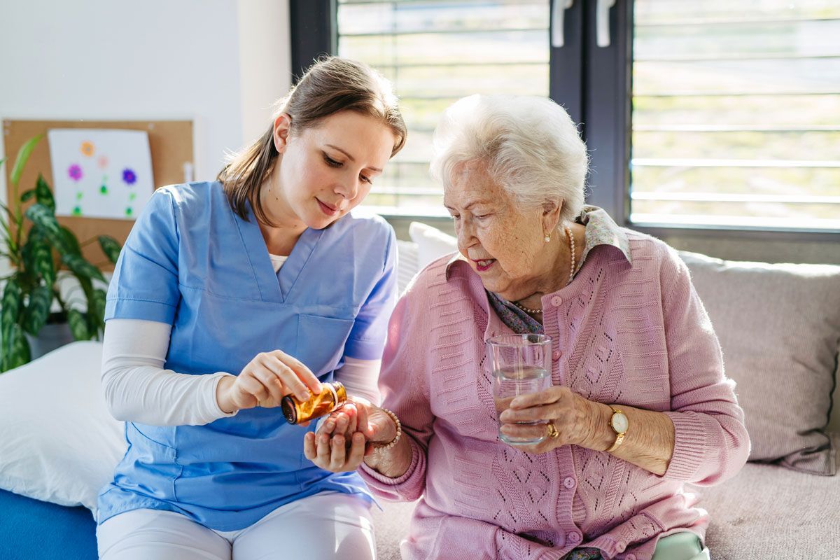 A caregiver in blue scrubs gives medication to an elderly woman in a pink sweater. Sunlight streams through a nearby window.