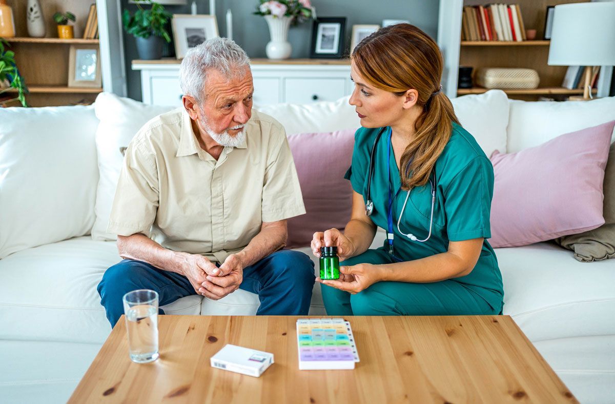 A nurse shows an elderly man a pill bottle and pill organizer while seated on a couch. The man looks intently at her.