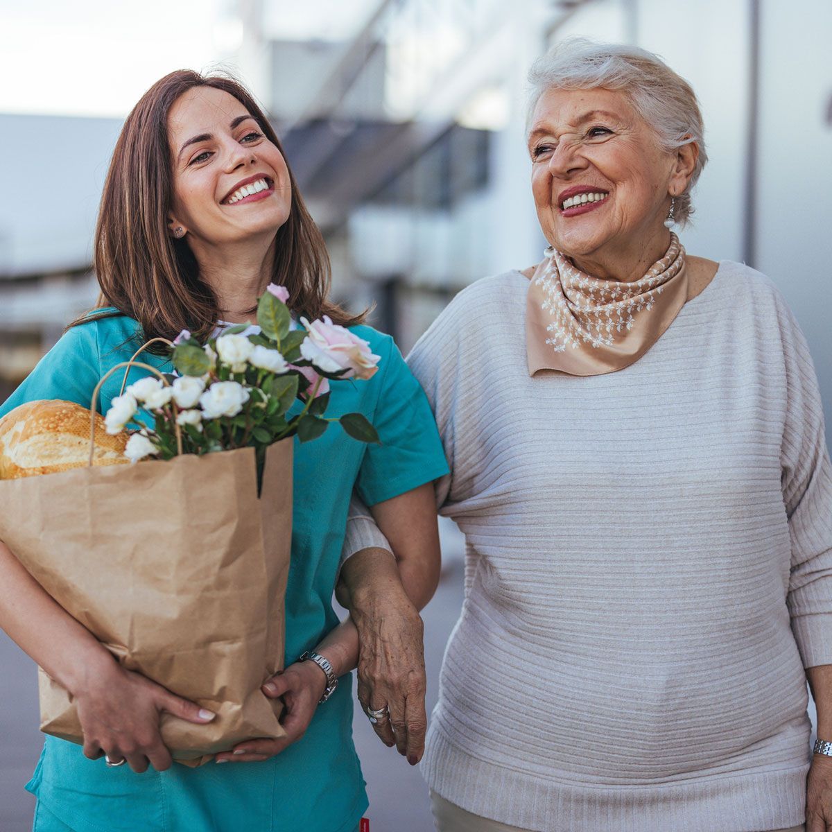 A smiling caregiver assists an elderly woman, holding groceries and flowers outside a building. Both women are smiling.