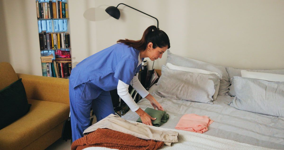 A woman in blue scrubs folds clothes on a bed. She stands in a bedroom with a bookshelf and a small sofa.
