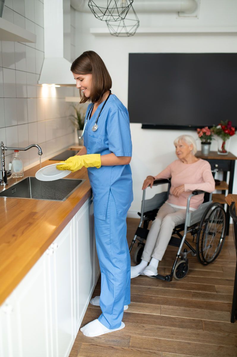 Caregiver washing dishes in a kitchen, while a senior woman sits in a wheelchair nearby. The caregiver wears blue scrubs and yellow gloves.