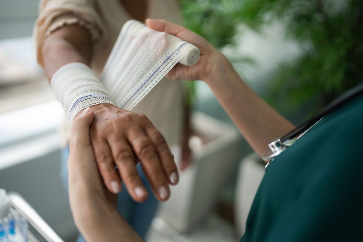 A medical professional bandages a person's wrist and forearm. Light-skinned hands are visible.