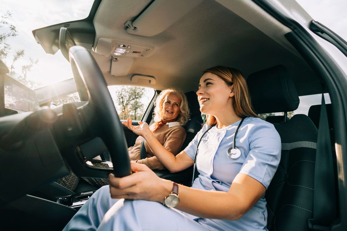 A young nurse drives a car with an older woman in the passenger seat, smiling. Both are inside a car on a sunny day.