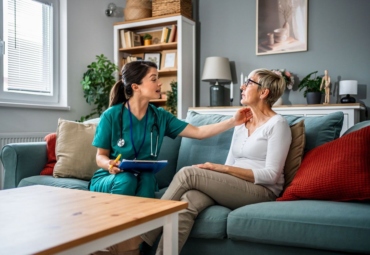 A medical professional in teal scrubs examines an older woman's neck in a living room. The woman sits on a blue sofa, looking toward the professional.