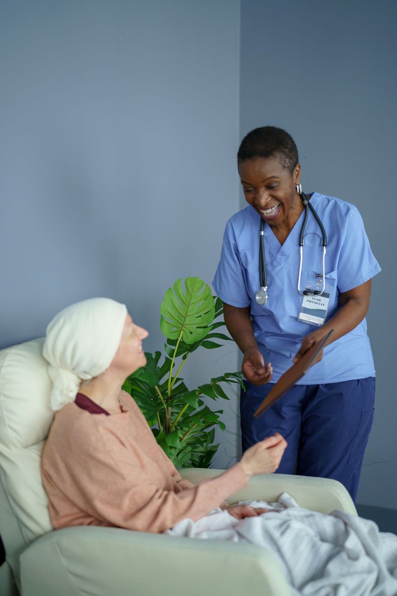 Nurse in blue scrubs smiles at a woman with a headscarf seated in a chair, indoors.