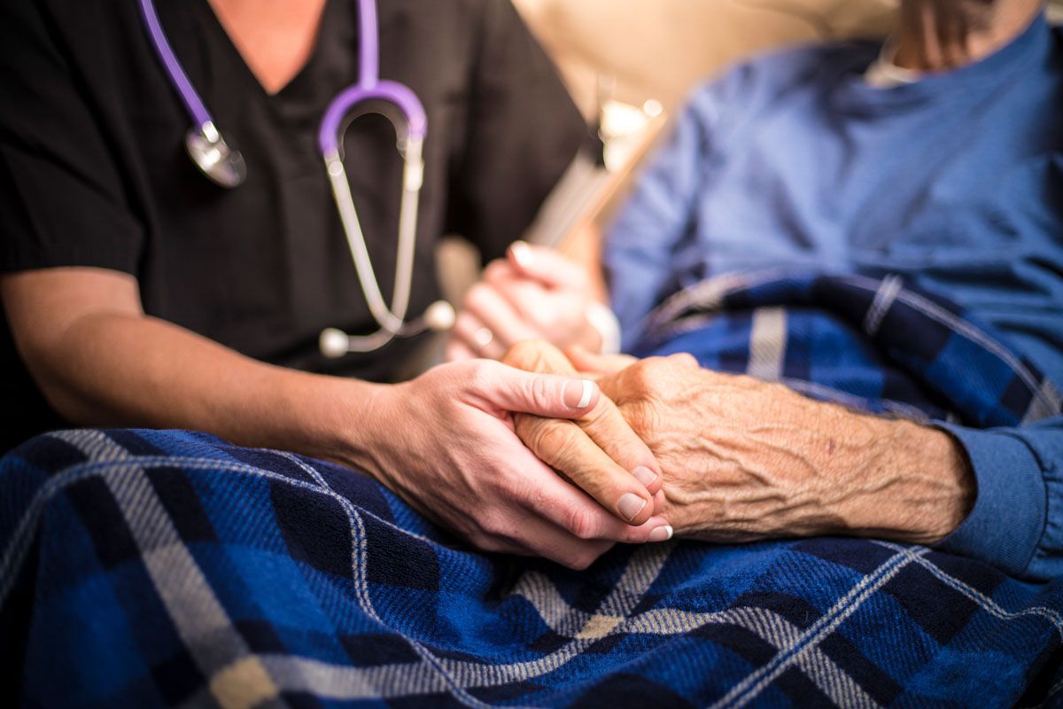 A healthcare worker holds the hand of an elderly person wearing a blue shirt and plaid blanket, likely offering comfort and support.