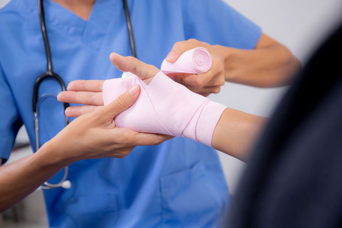 A medical professional in blue scrubs wrapping a pink bandage around a patient's hand in a clinical setting.