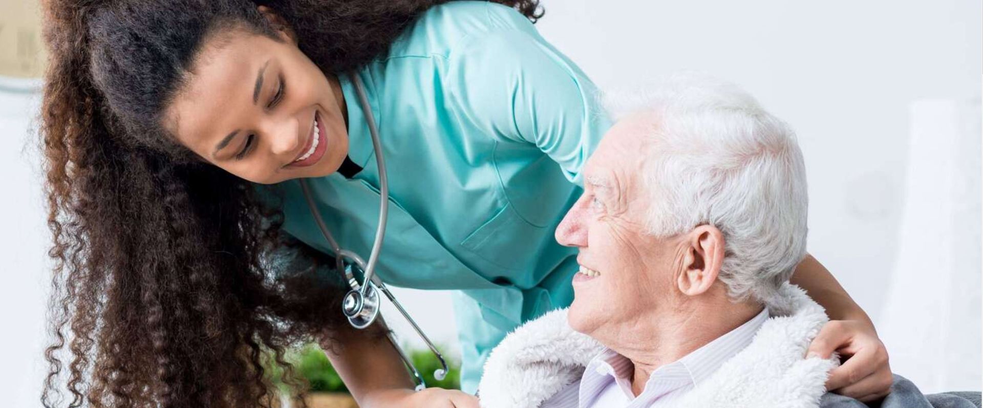 A smiling nurse in teal scrubs comforts an elderly man with white hair, indoors.