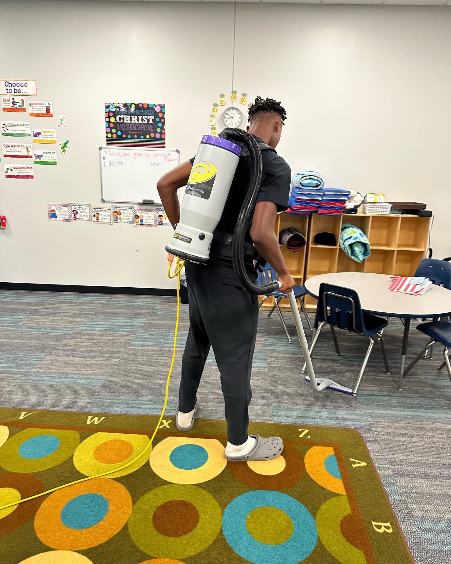A man is cleaning a rug in a classroom with a backpack vacuum cleaner.
