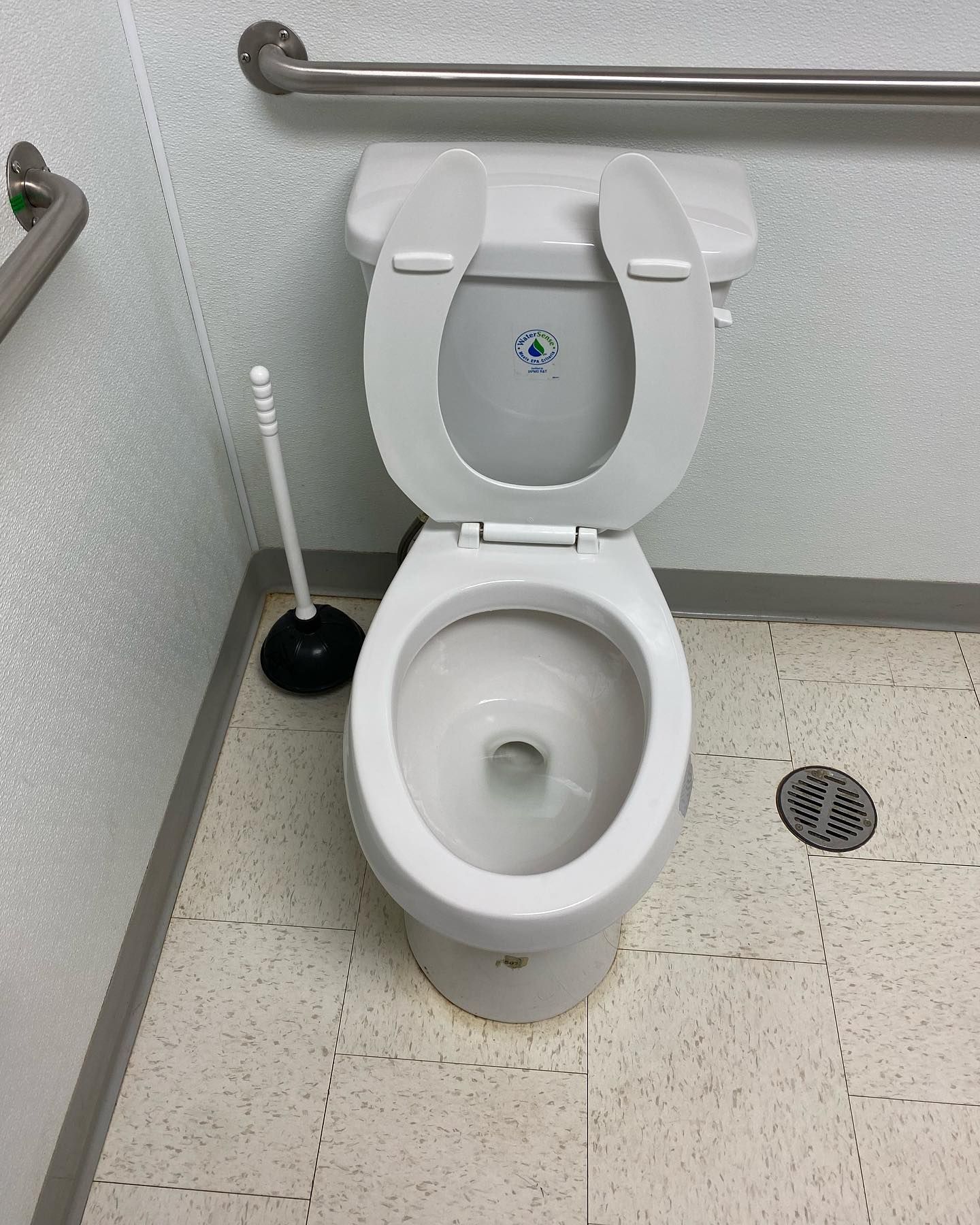 A toilet with the seat up and a plunger on the floor