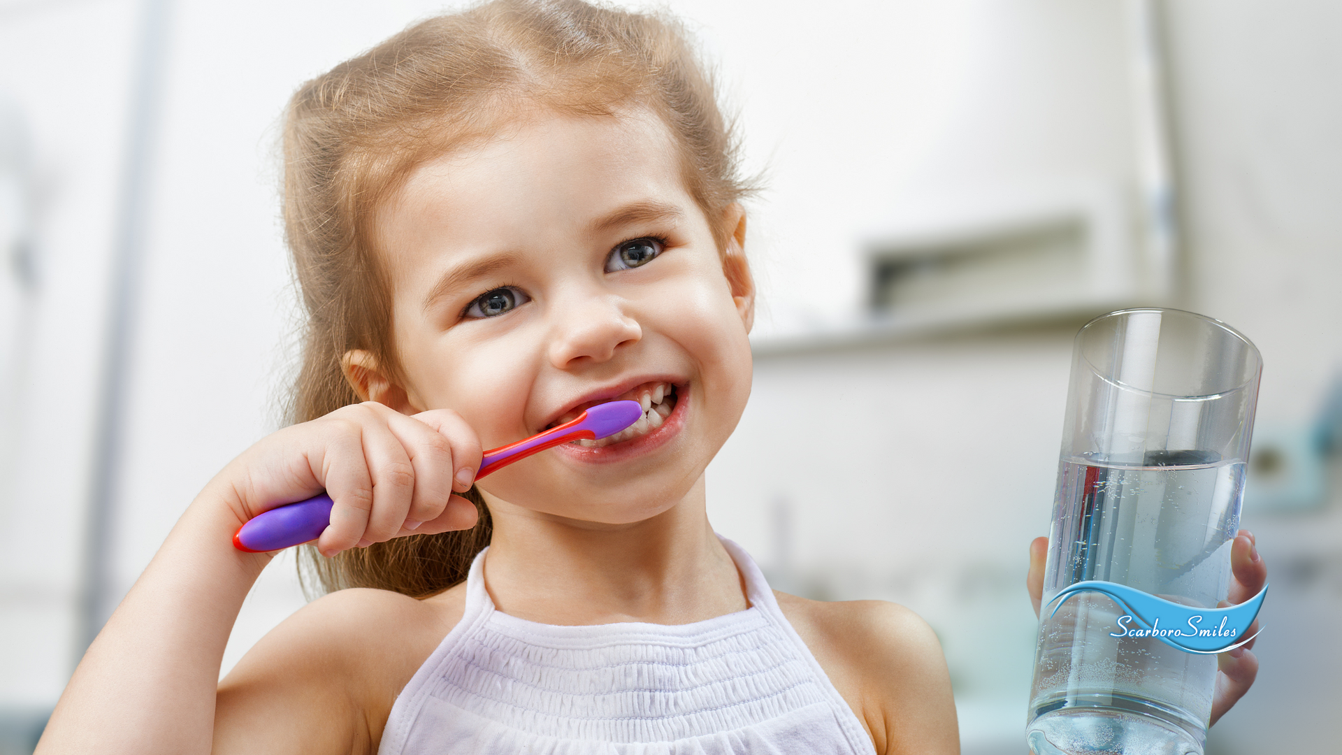 A little girl is brushing her teeth while holding a glass of water.