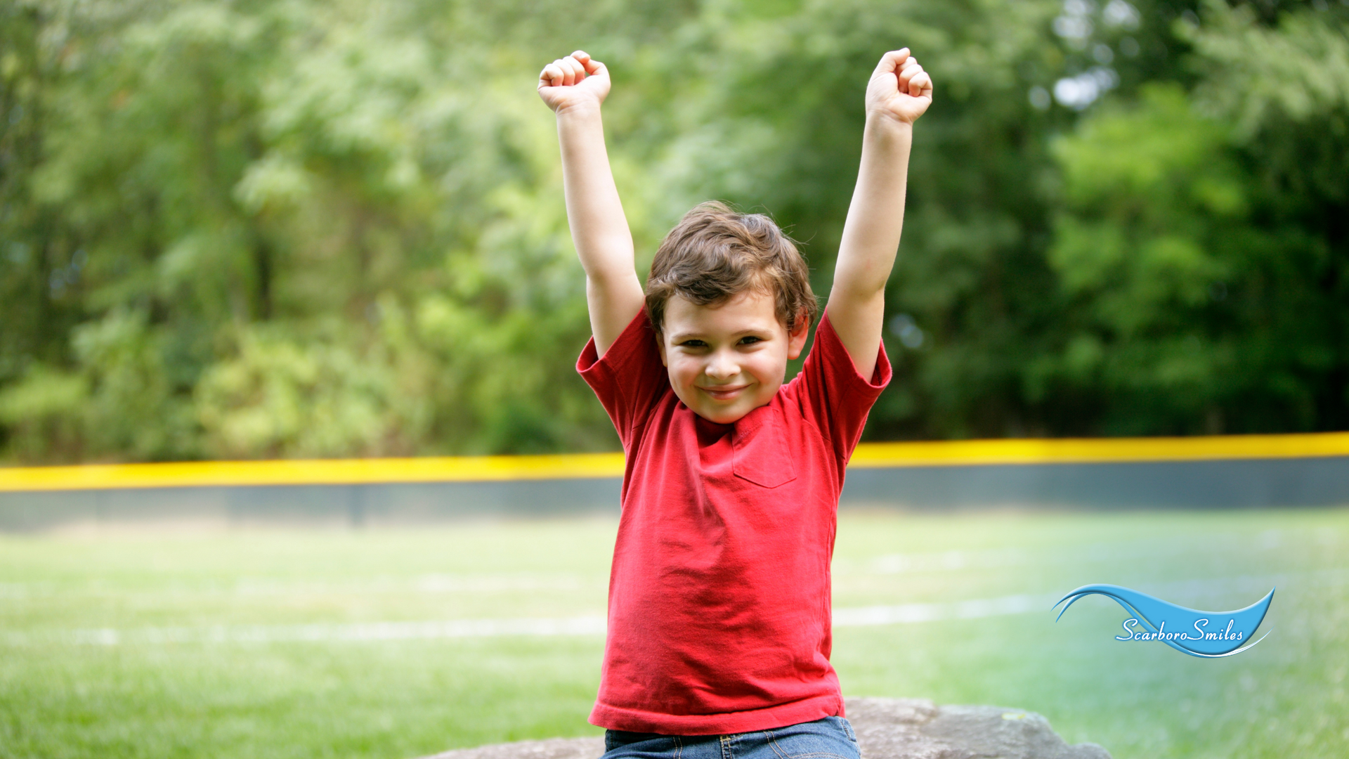 A young boy in a red shirt is sitting on a rock with his arms in the air.