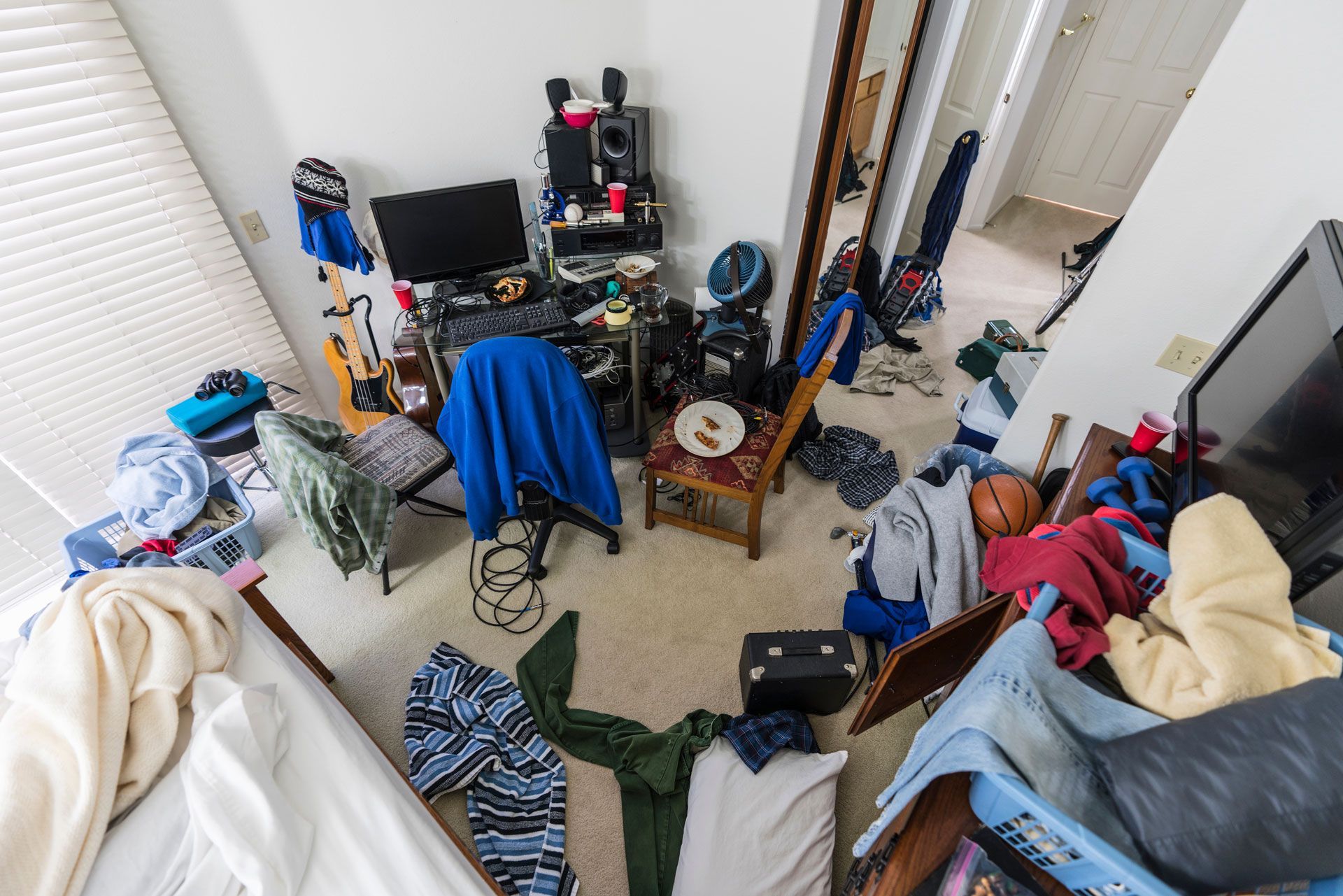 A bedroom with a lot of clothes on the floor and a computer desk.