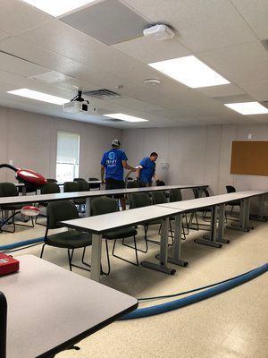 Two men are cleaning a classroom with tables and chairs.