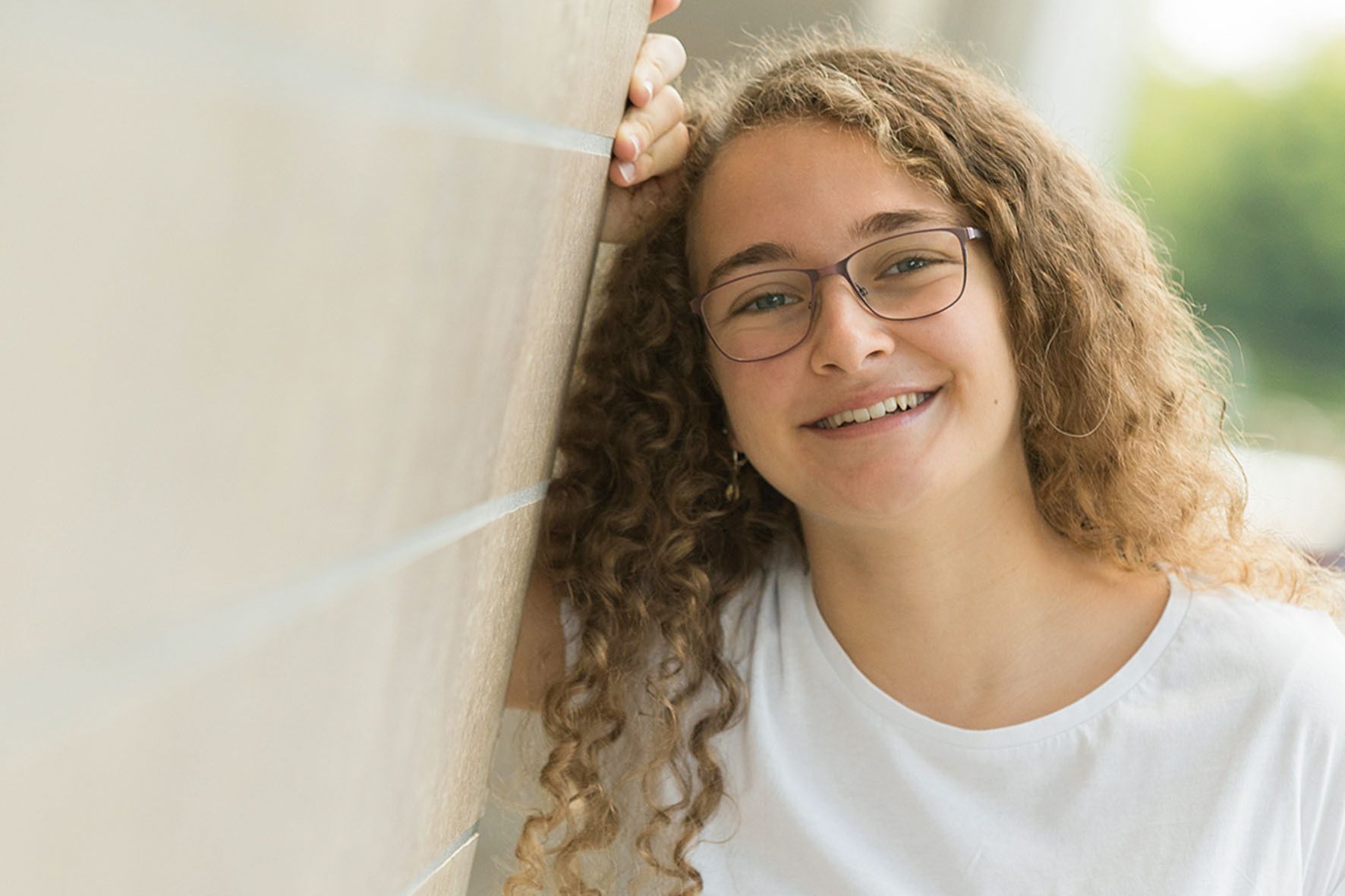 Eine junge Frau mit lockigem Haar und Brille lehnt an einer Wand und lächelt.