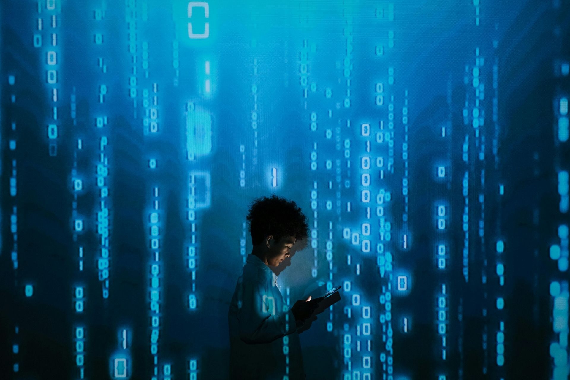 Person in digital space, illuminated by blue binary code; holding a tablet, looking thoughtful.