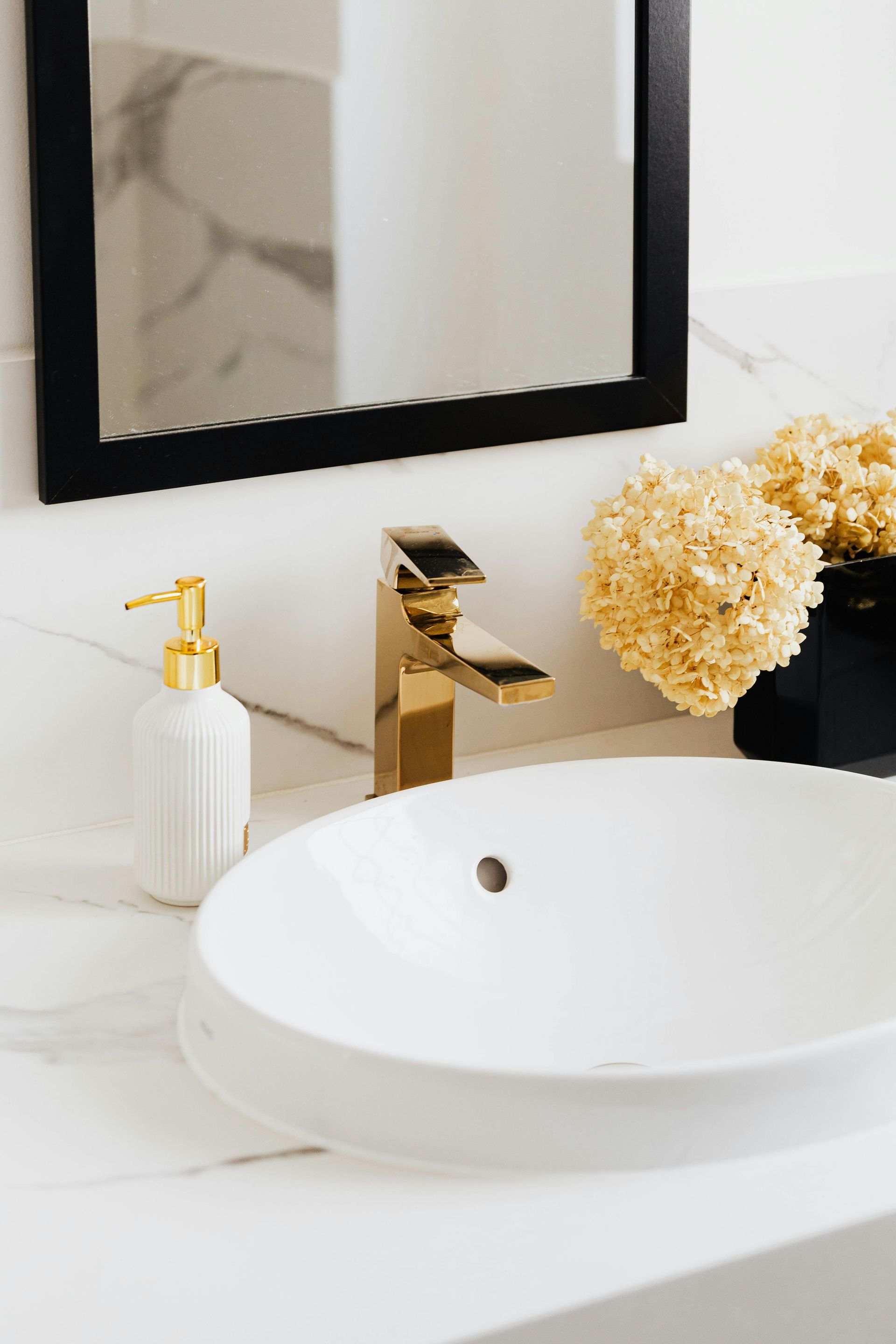 A bathroom with a sink , mirror , soap dispenser and flowers.