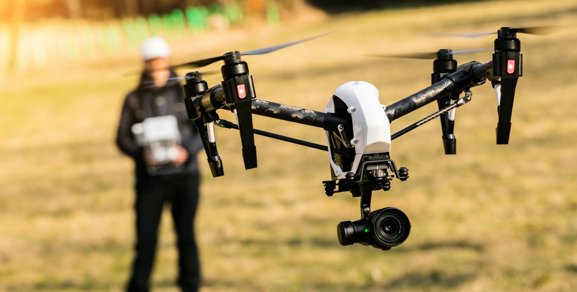 Drone in flight with a person in background holding a remote. Outdoor, sunny setting.