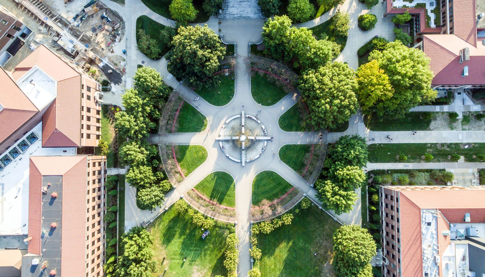 Aerial view of a garden with paths radiating from a central fountain, surrounded by trees and buildings.