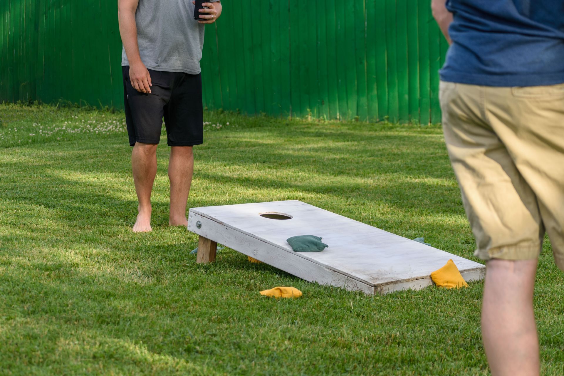 Two people playing cornhole on a grassy lawn with a green fence in the background.