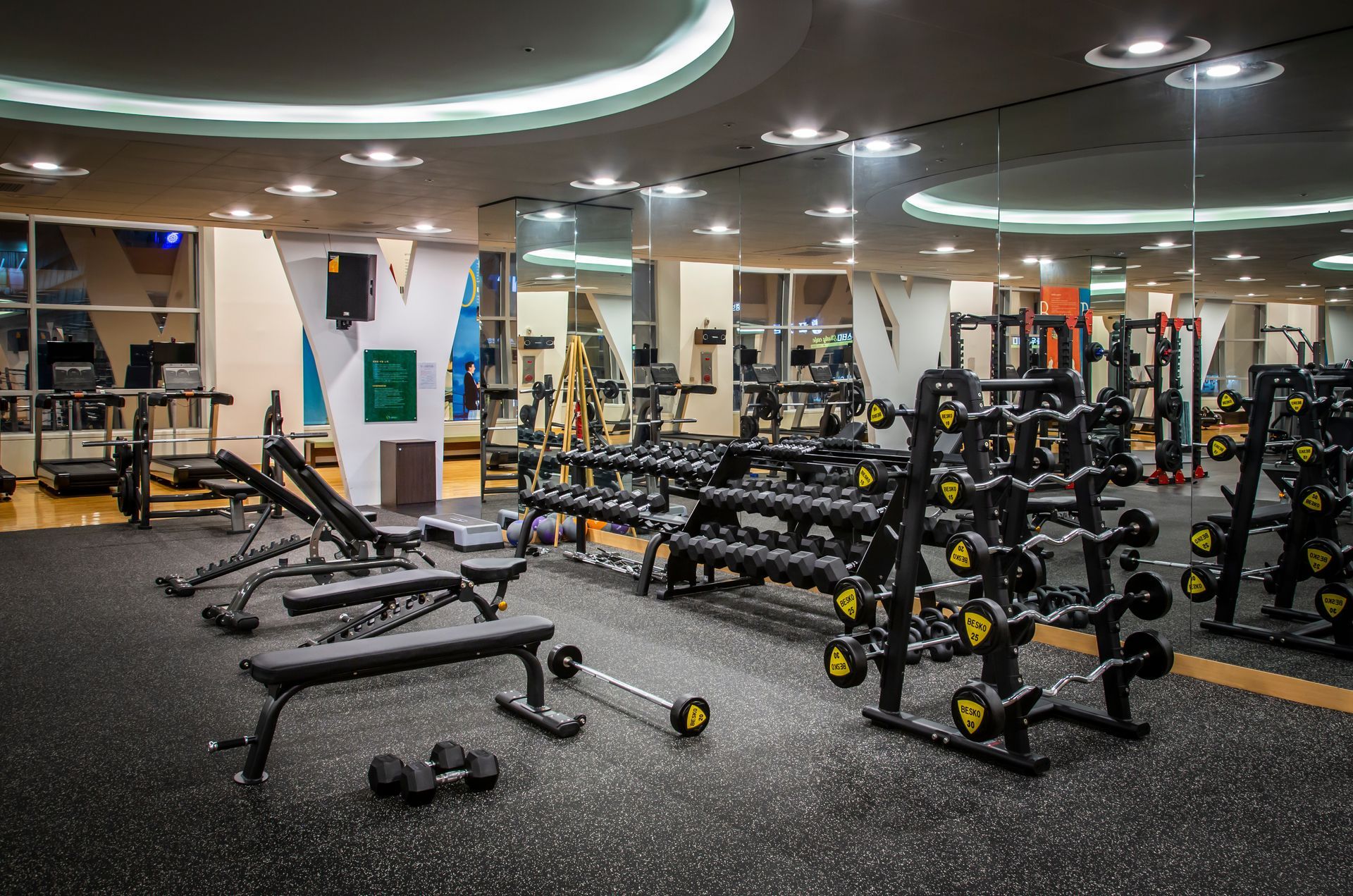 Gym interior with weight racks, benches, and exercise machines under bright lighting.
