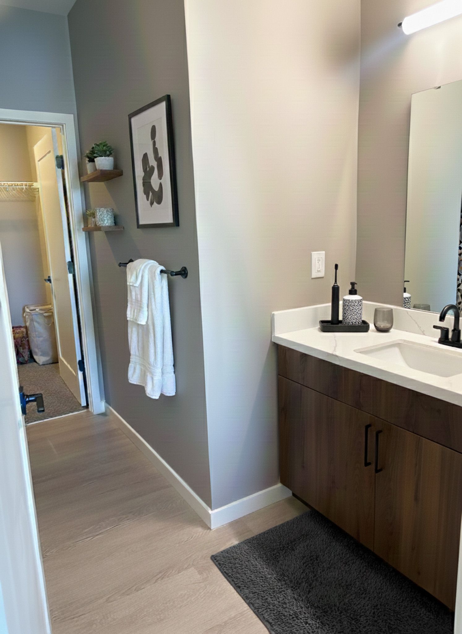 Bathroom with wooden vanity, white countertop, and gray walls. Towel rack and art on wall, open closet visible.