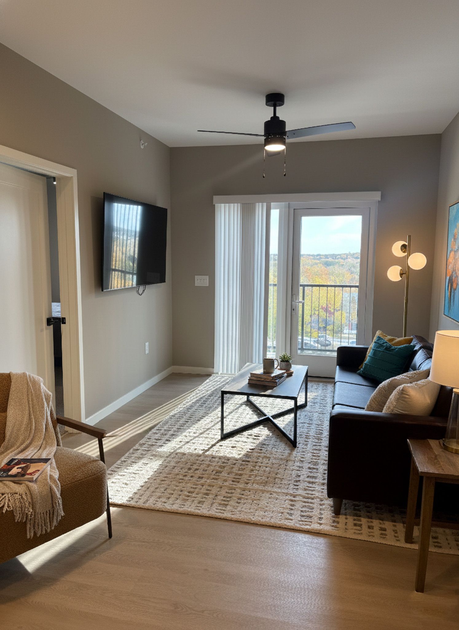 Living room with couch, TV, rug, and balcony access. Beige walls and hardwood floors. Sunlight streams through the window.