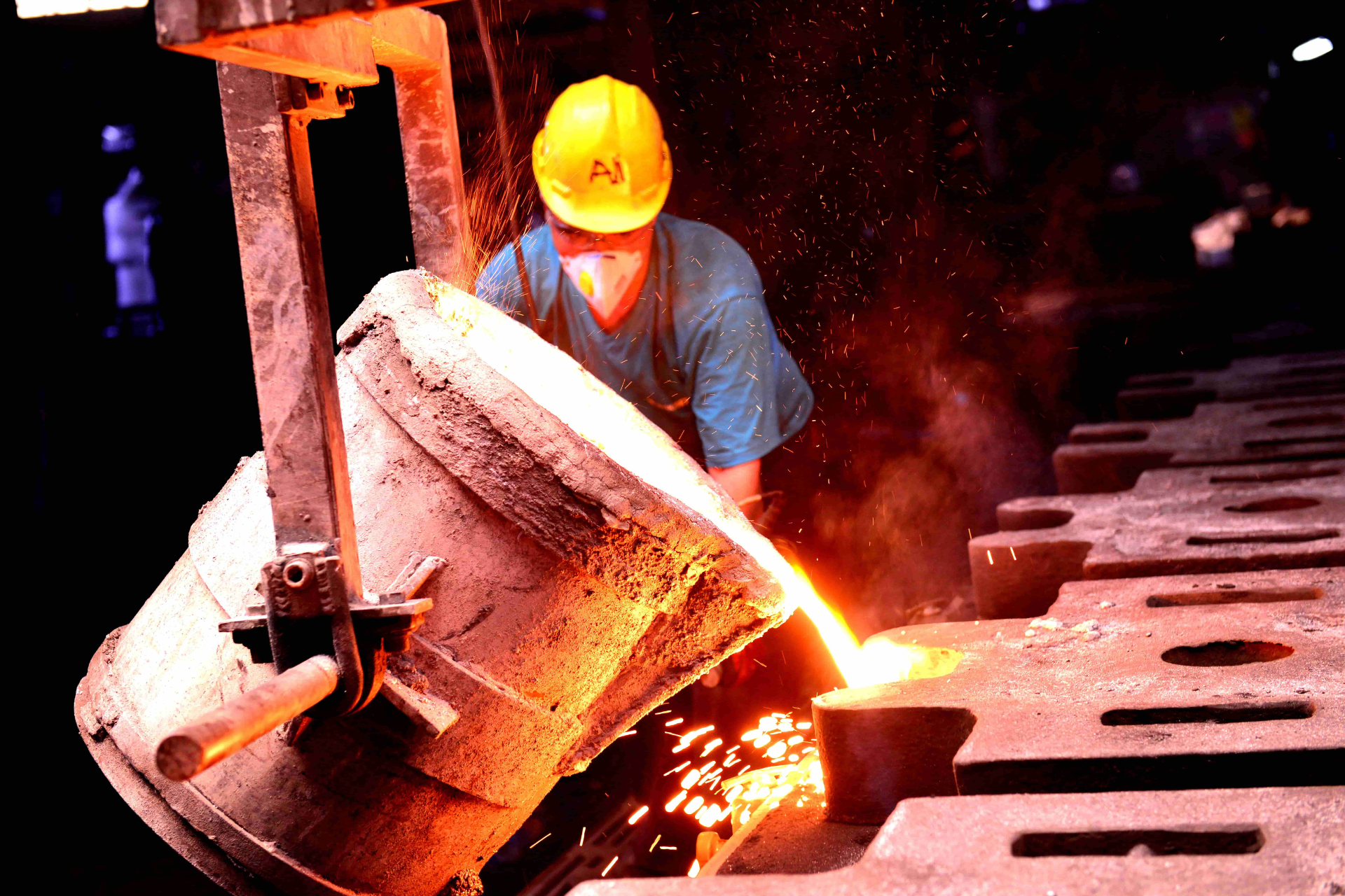 A worker wearing a yellow hard hat and face mask pours molten metal from a large crucible into molds