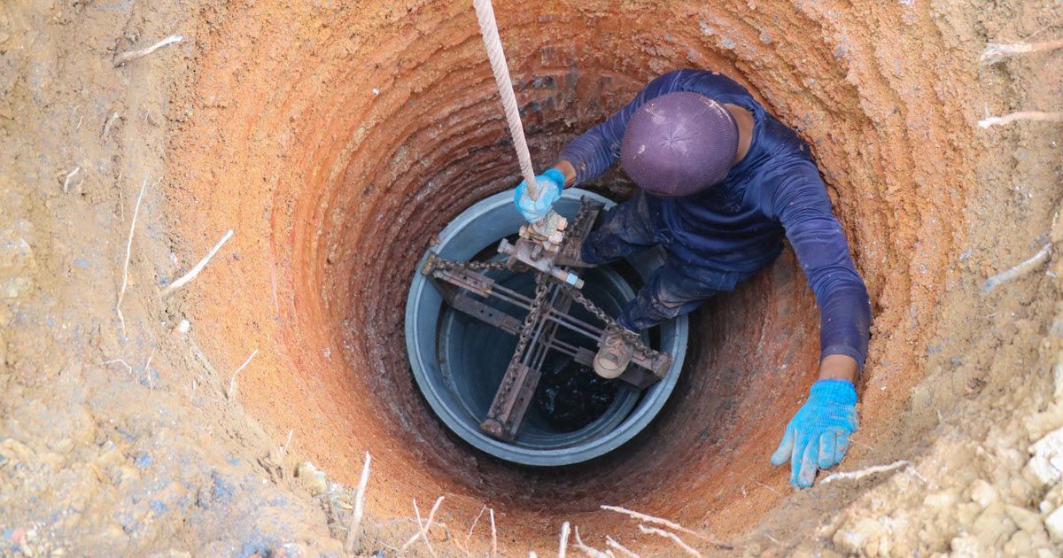 A person in a well, using a winch to pull up a bucket. The well is earthen, and the person is wearing blue gloves and a purple helmet.Hydrogeology Projects - NYC, Manhattan, The Boroughs, Nassau County, Suffolk County, Long Island, and New Jersey - Kagaoan Engineering