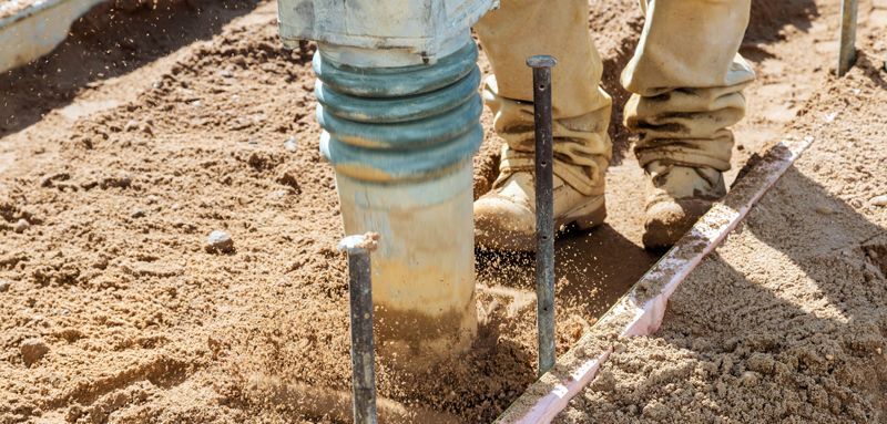A construction worker uses a jumping jack to compact sand. The machine is beige and teal, and the worker wears tan work boots. Techniques for Soil Stabilization: Enhancing Ground Strength