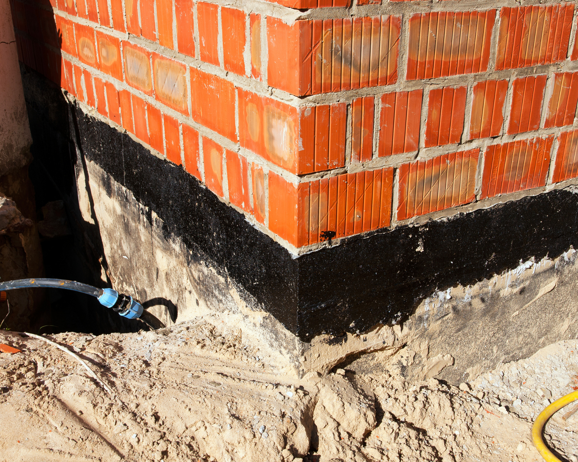 Brick building corner with black waterproofing layer applied to the foundation, surrounded by sand.