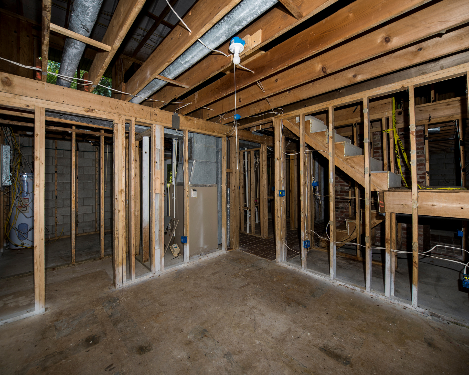 Unfinished basement with exposed wood framing, plumbing, and electrical wiring. A staircase is visible. The floor is concrete.