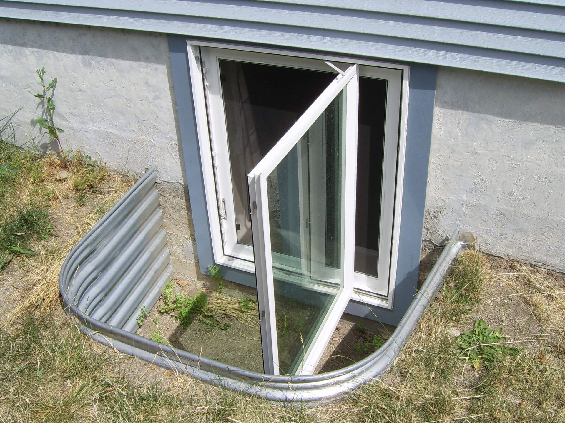 A partially open basement window with a metal window well, set in a gray concrete wall and grass.
