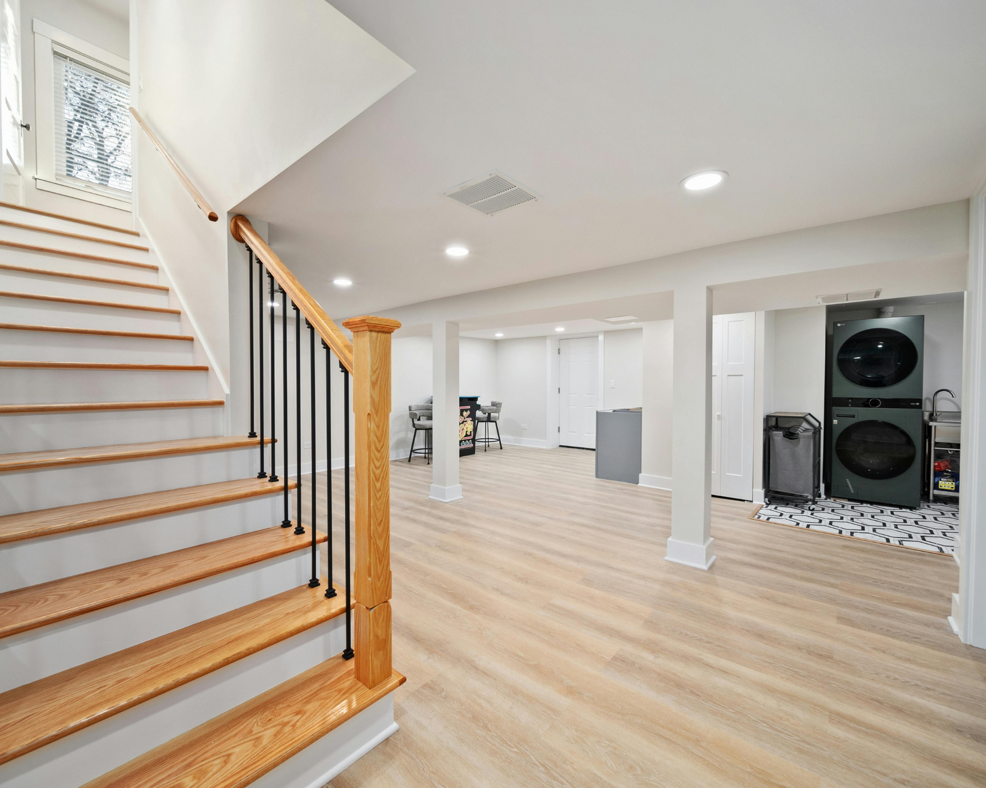 A bright basement with a staircase, laundry machines, and light wood flooring. The walls and columns are white.