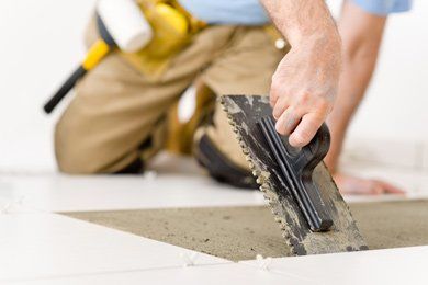 A person installing tiles on a floor, using a trowel to spread mortar. They are kneeling, wearing work clothes.