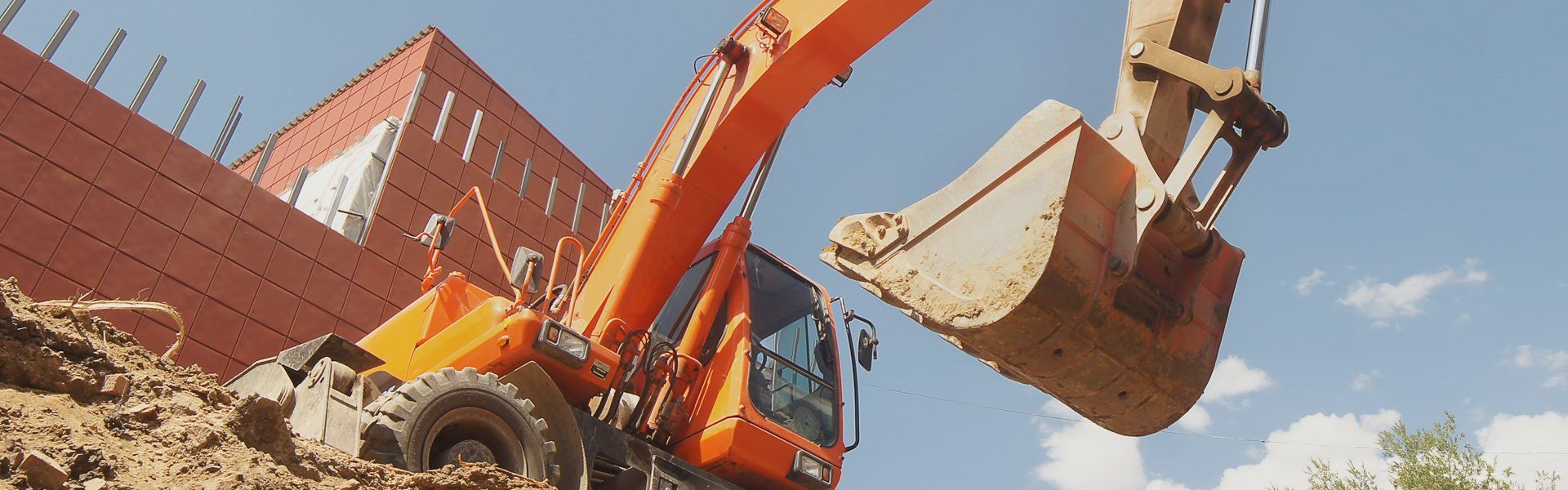 An orange excavator with a large bucket is in action, digging soil at a construction site with a brick building in the background.