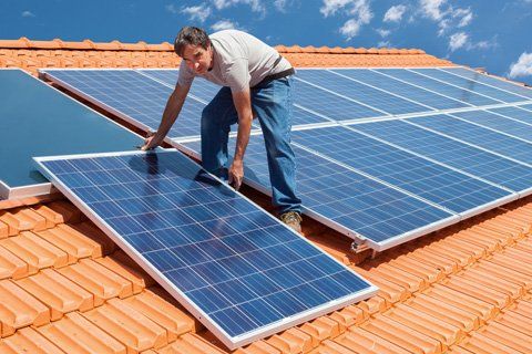 A man installs a solar panel on a red tile roof under a blue sky.