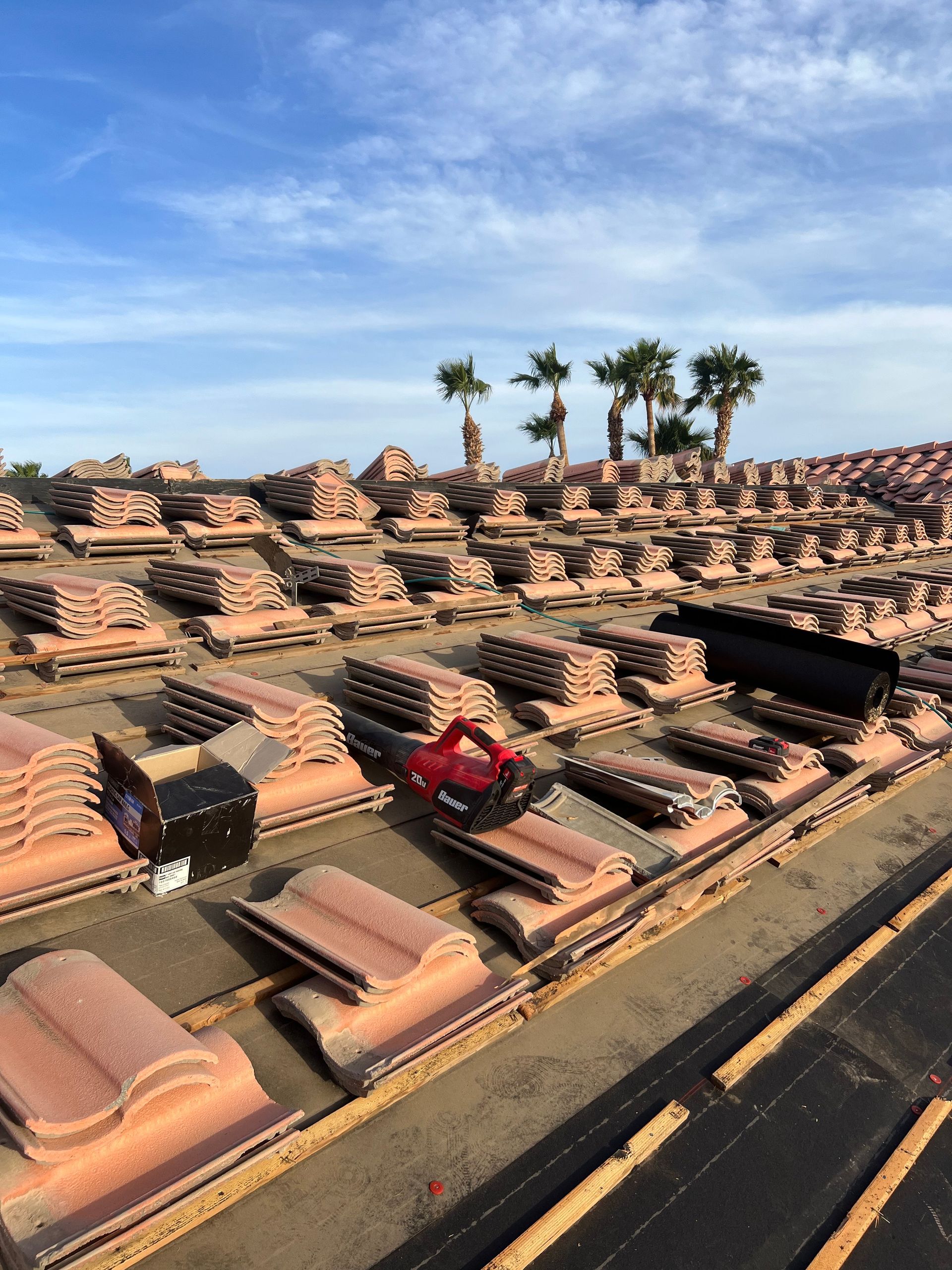 Rooftop with stacks of clay tiles, a power saw, and palm trees under a blue sky.