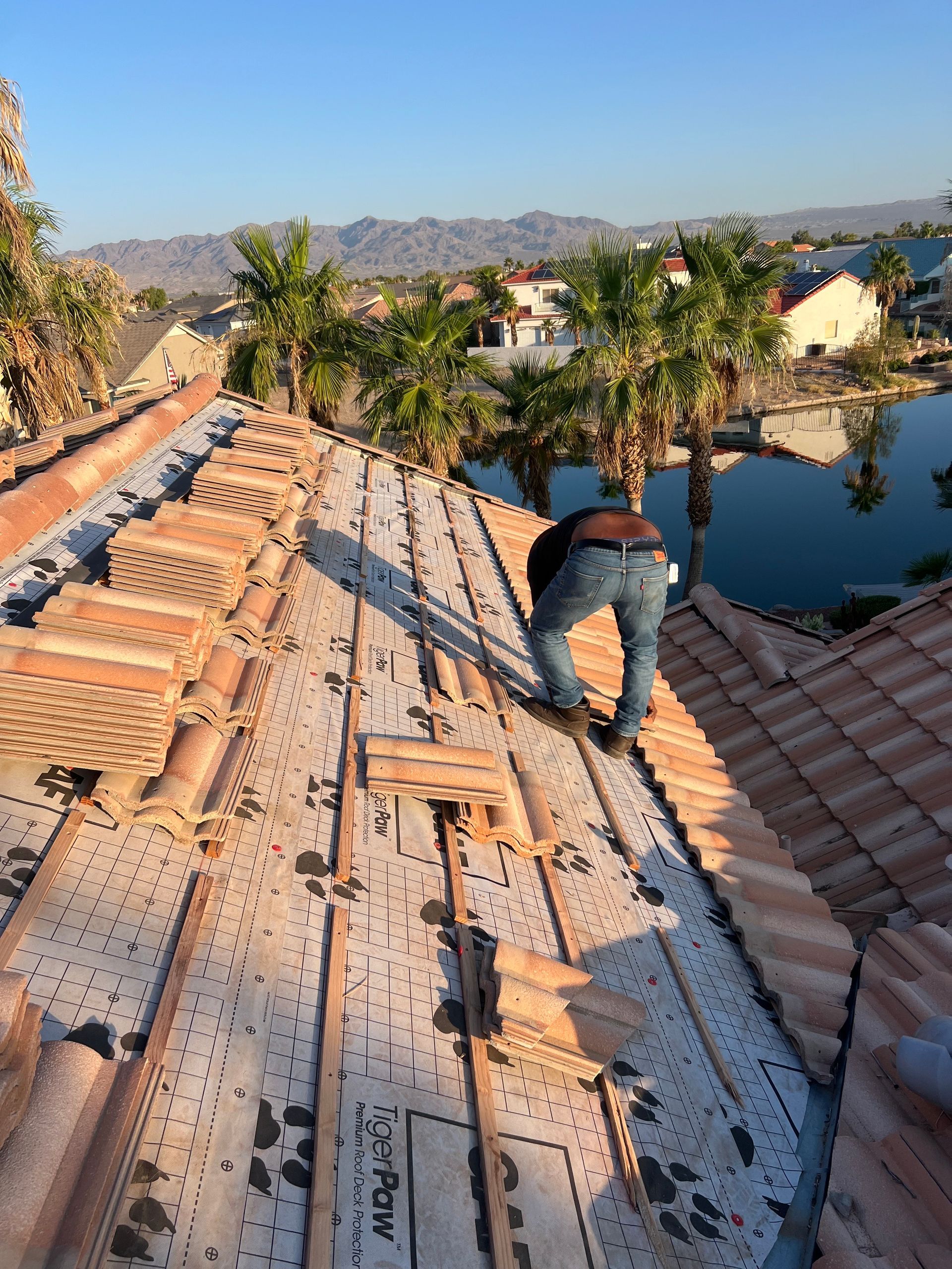 Man on a rooftop installing clay roof tiles. Sunny day, water and palm trees visible in the background.