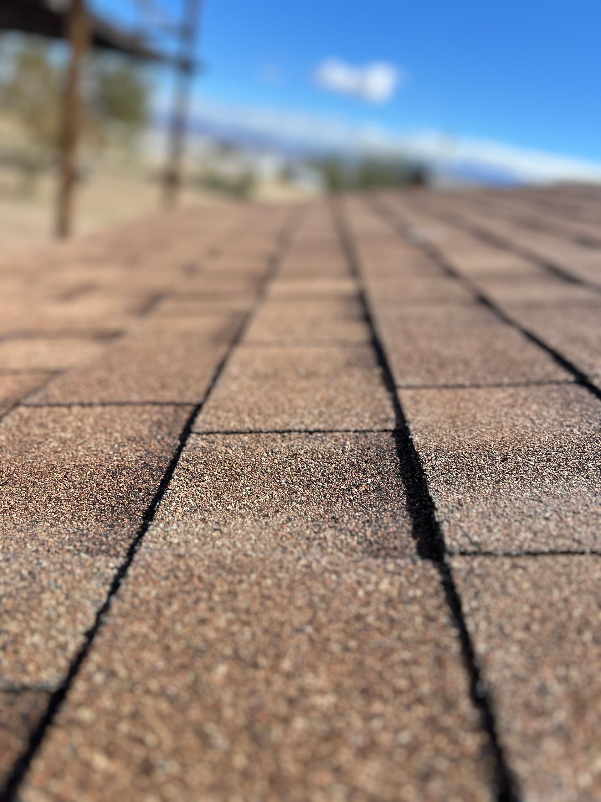 Close-up of brown asphalt shingles on a roof, with a blurred blue sky and desert landscape in the background.