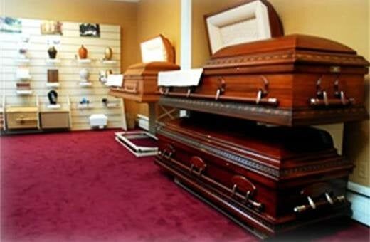 Funeral home showroom with three wooden caskets displayed on a red carpet, with urns and other merchandise on shelves in the background.