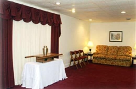 Funeral home interior: display table with urn, red carpet, curtains, sofa, chairs.