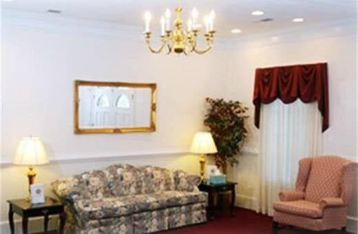 Funeral home waiting area with sofa, chair, lamps, and decorative window drapes.