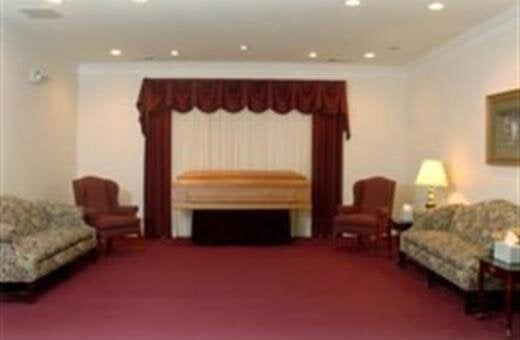 Funeral home interior, burgundy carpet, tan casket, maroon drape, floral couches, chairs, and lamps.