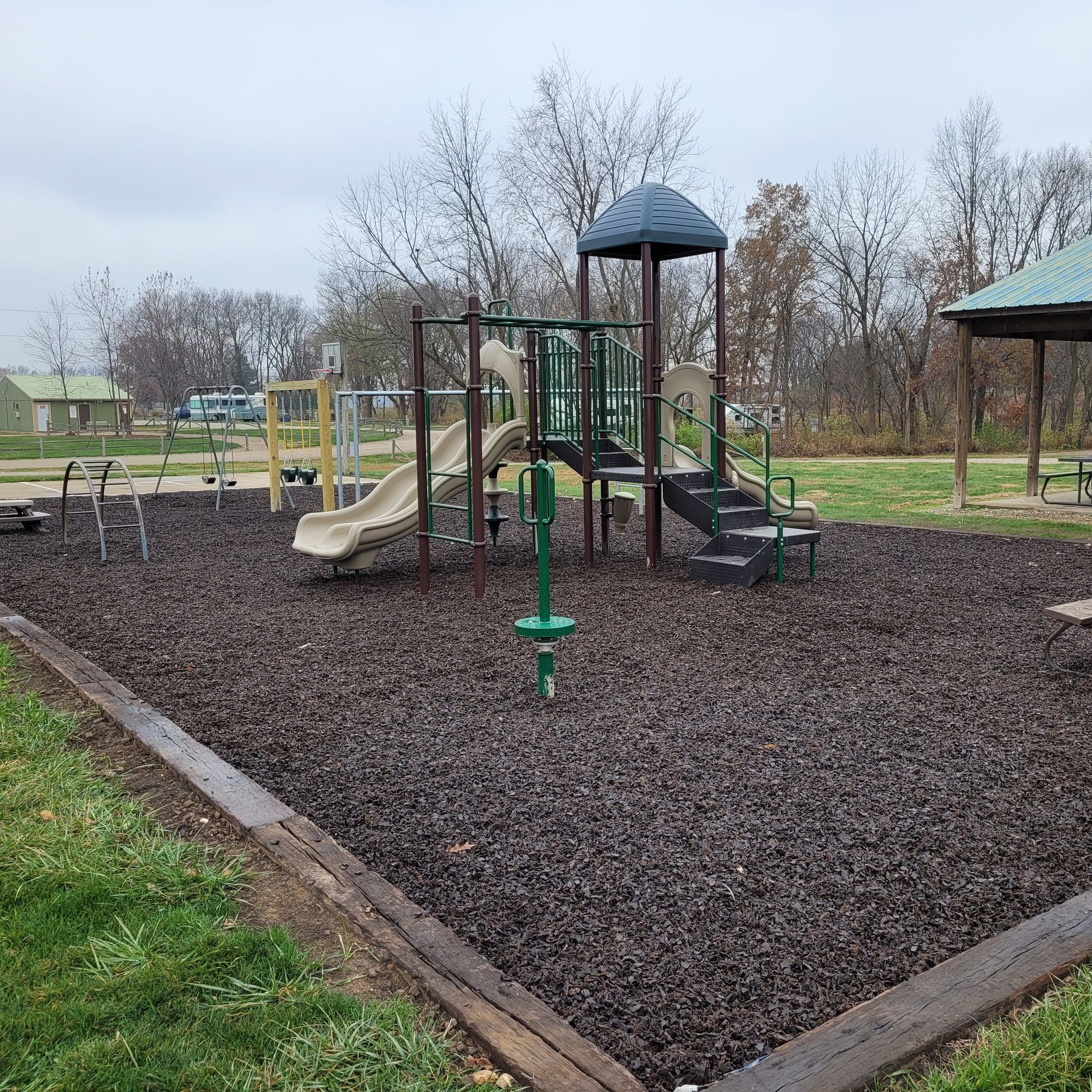 A playground with a slide , stairs , and benches in a park on a cloudy day.