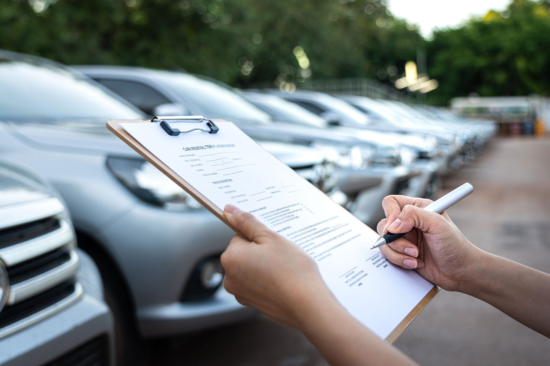 Person writing on a clipboard, inspecting cars parked in a row outdoors.