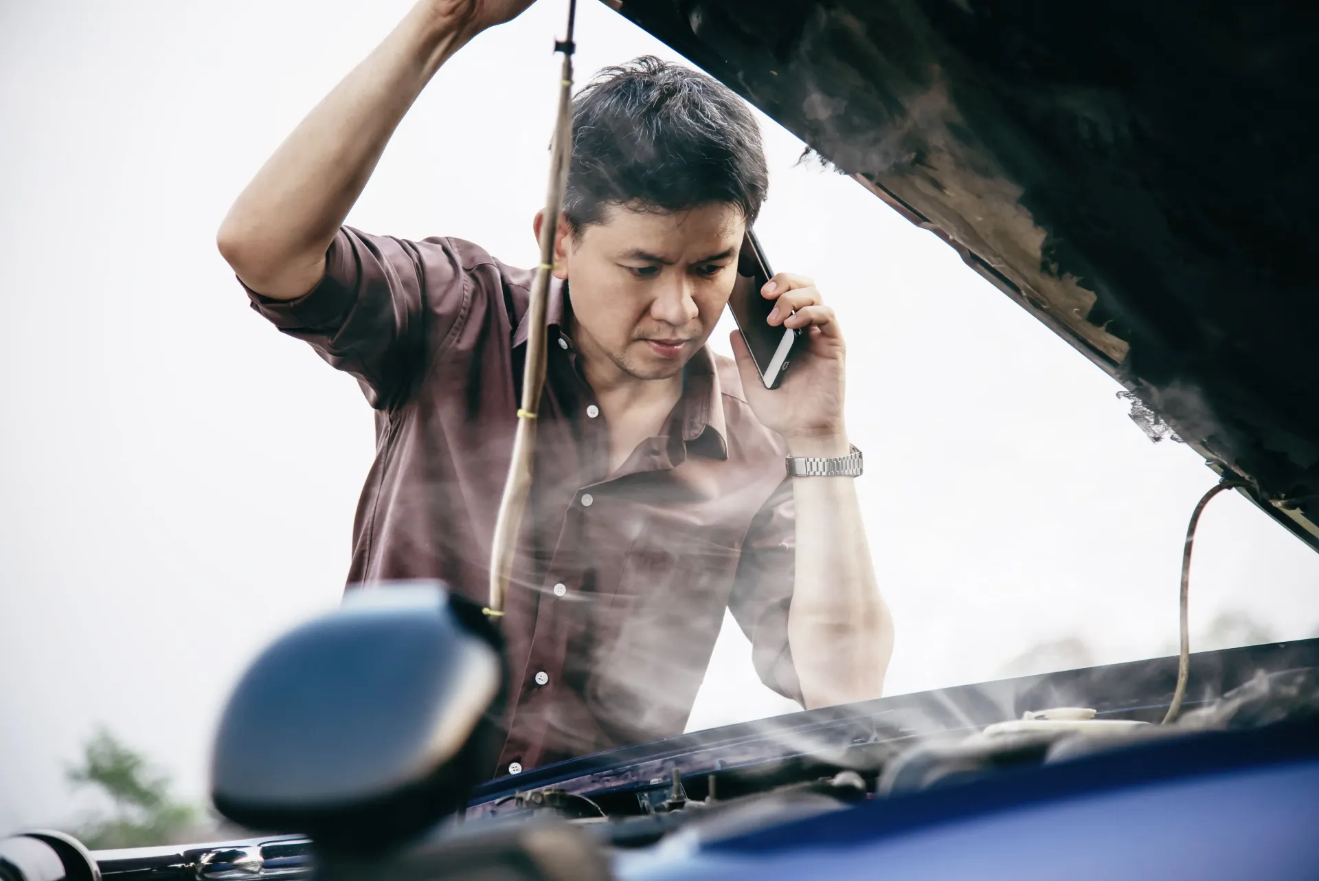 A worried man looks under his car’s hood while talking on the phone as smoke rises from the engine, 