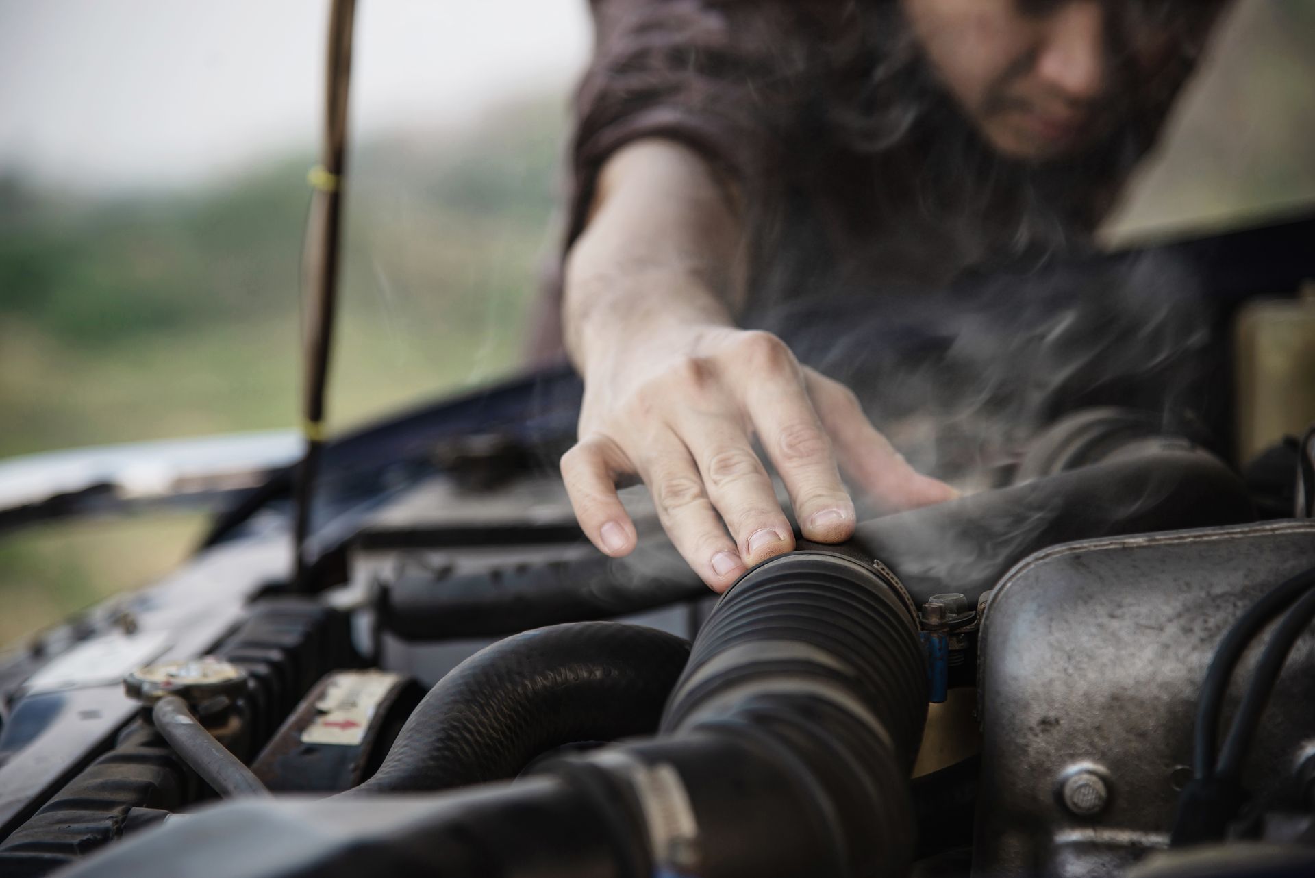 A person checking under the hood of a smoking car engine, indicating a possible overheating issue.
