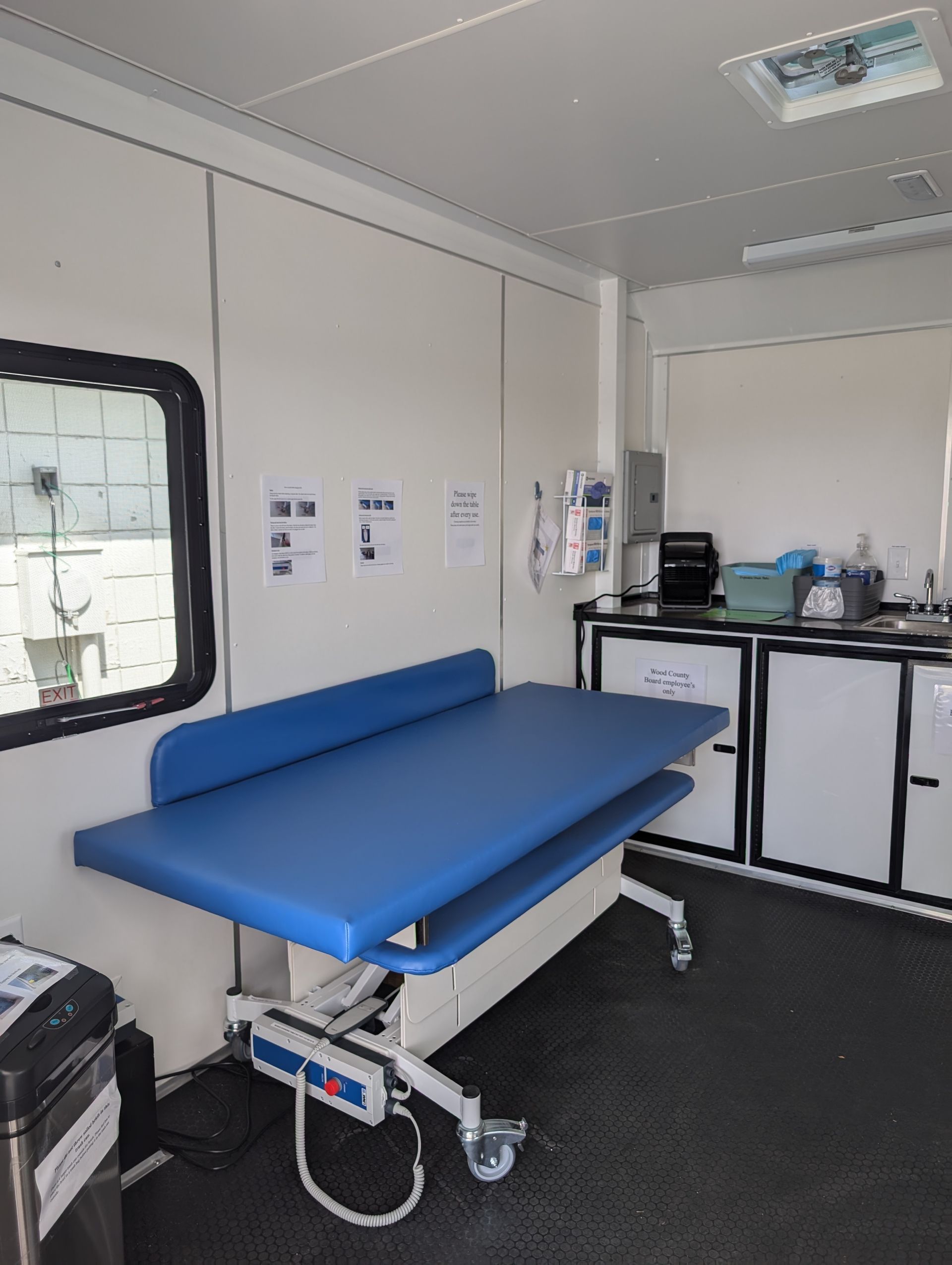 Interior of a universal changing room. Features an examination table with blue pad, white cabinets, and a black trash can.