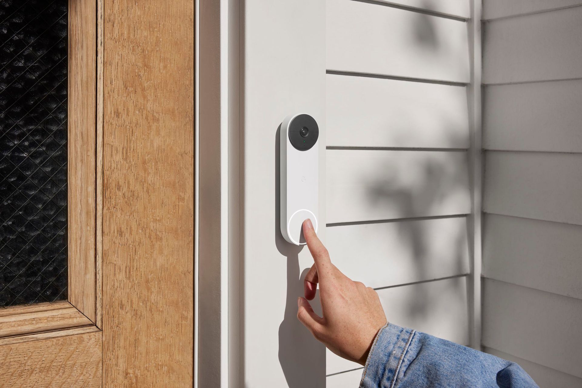 A person's hand pressing a white video doorbell on a white house, next to a wooden door.